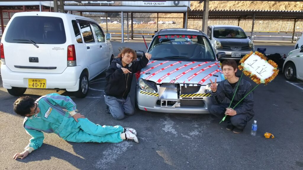 カーサービスオカダ代表がサーキットイベントで入賞した車両と仲間と写る、車への情熱と整備経験を伝える画像